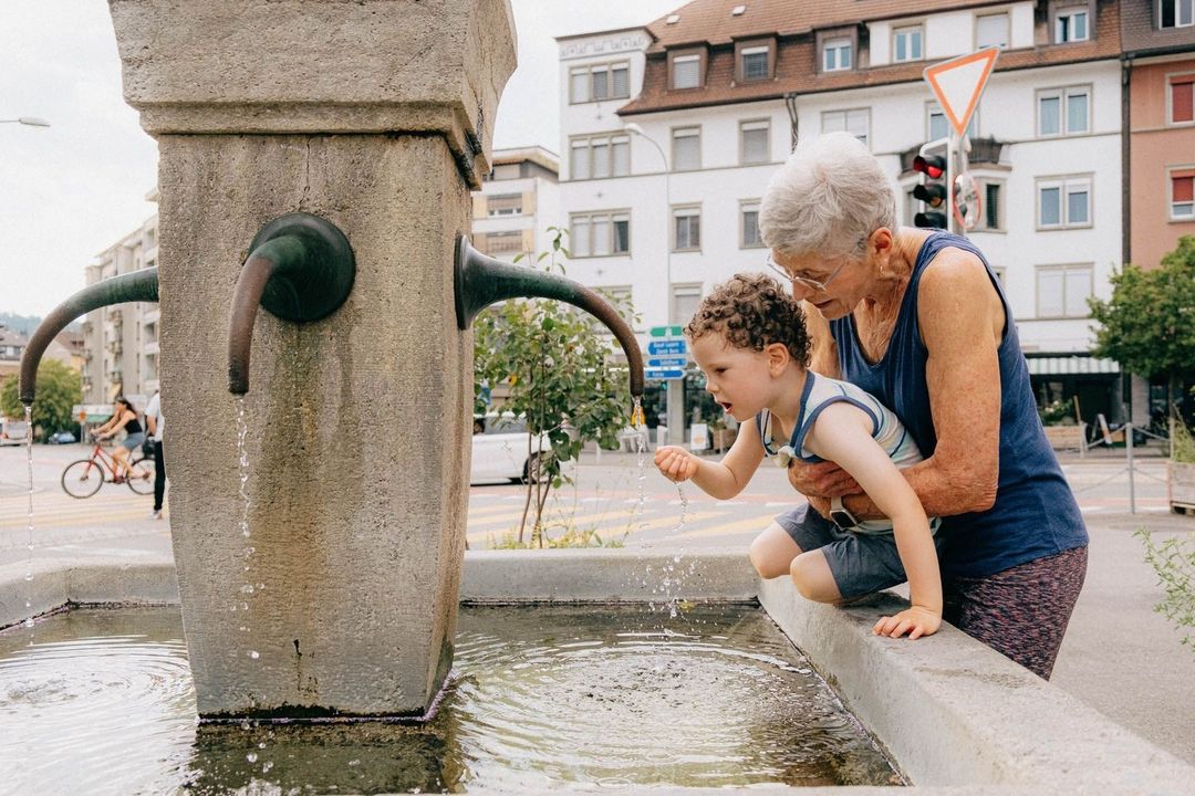 Outdoor shot of an elderly woman and a child interacting with a stone water fountain on a city street in daylight, with buildings and trees in view.