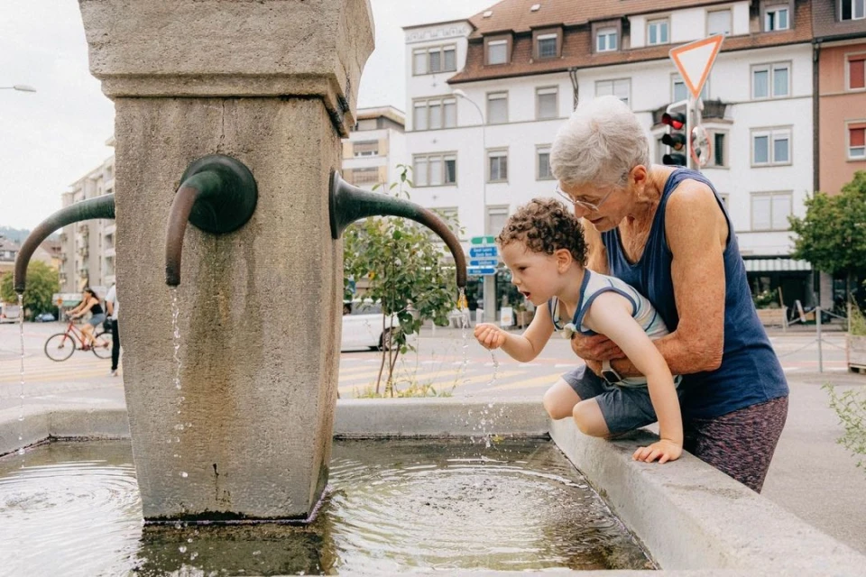 Outdoor shot of an elderly woman and a child interacting with a stone water fountain on a city street in daylight, with buildings and trees in view.