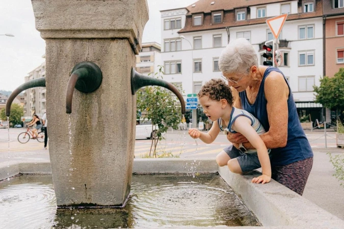 Outdoor shot of an elderly woman and a child interacting with a stone water fountain on a city street in daylight, with buildings and trees in view.