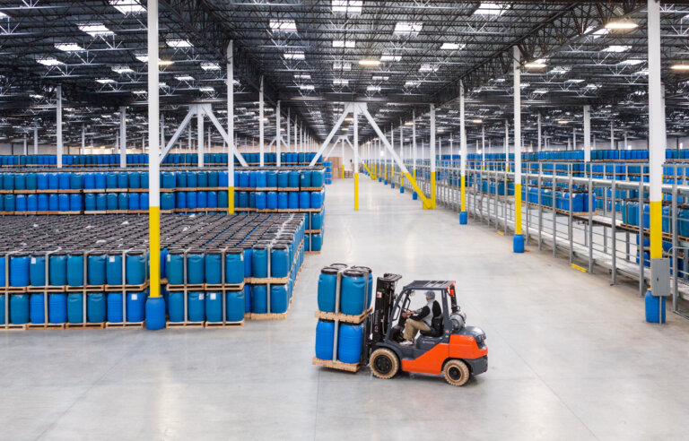 Large indoor warehouse with tall stacks of blue chemical barrels and a worker operating an orange forklift to move product.