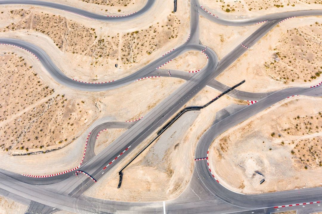 Aerial view of a winding asphalt race track with red and white safety barriers, surrounded by desert landscape in daylight.