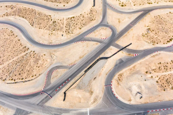 Aerial view of a winding asphalt race track with red and white safety barriers, surrounded by desert landscape in daylight.
