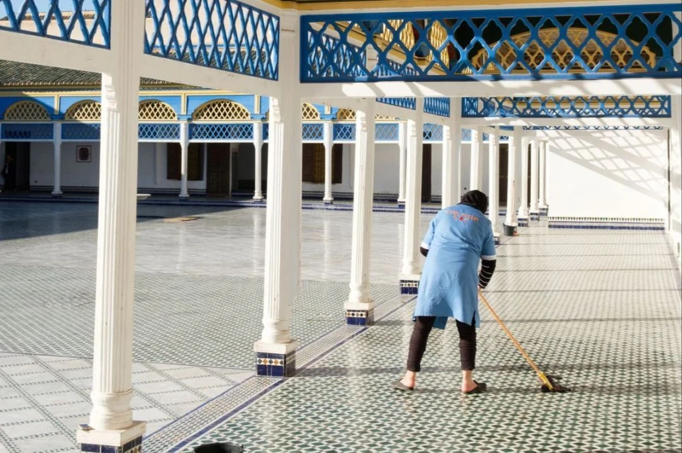 Woman sweeps tiled floor in ornate colonnaded courtyard. Architectural details include white columns and blue decorative lattice.