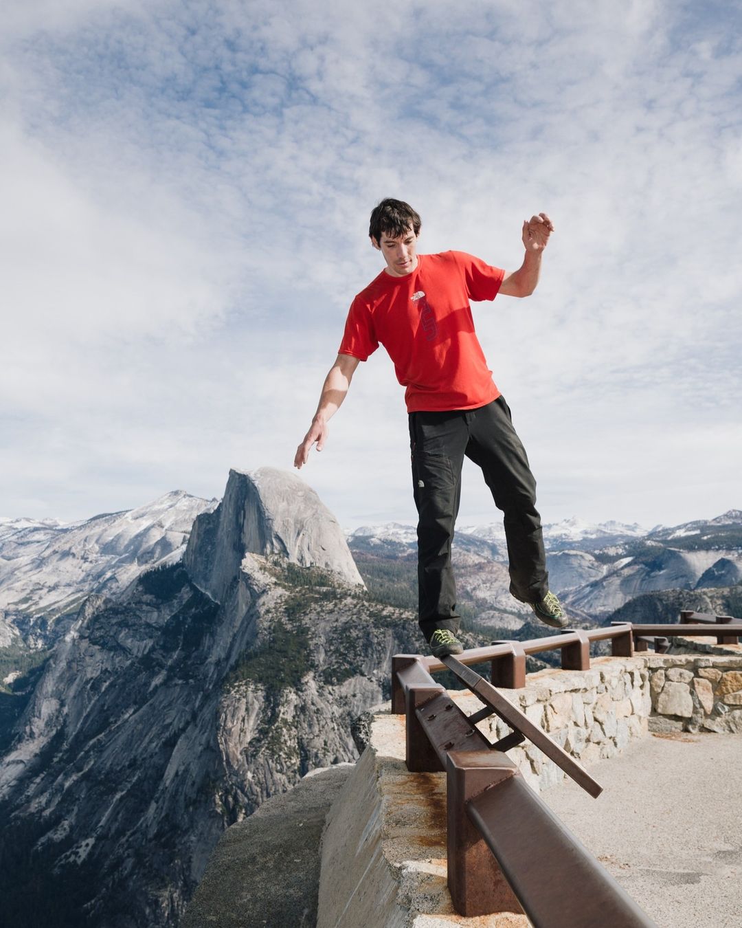 Athletic man balancing on a stone wall with a mountain view backdrop, captured on a sunny day with clear blue skies.