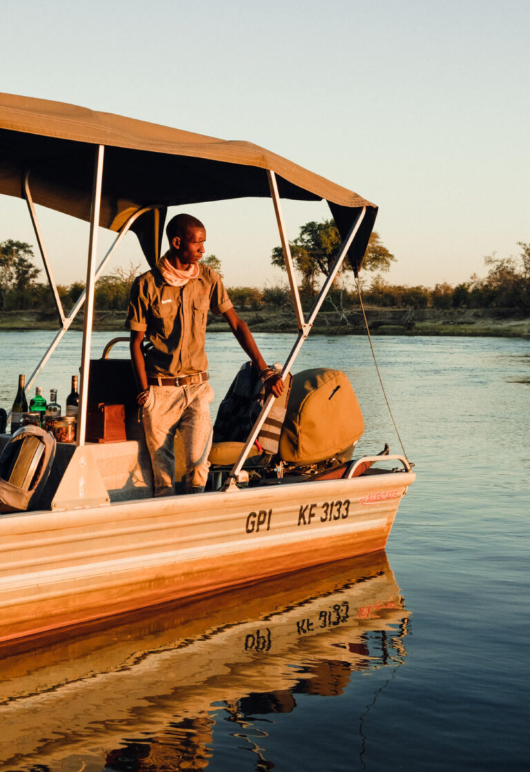 Man on a small boat with bottles on a table in a river; the boat's reflection shows in the water on a sunny day.
