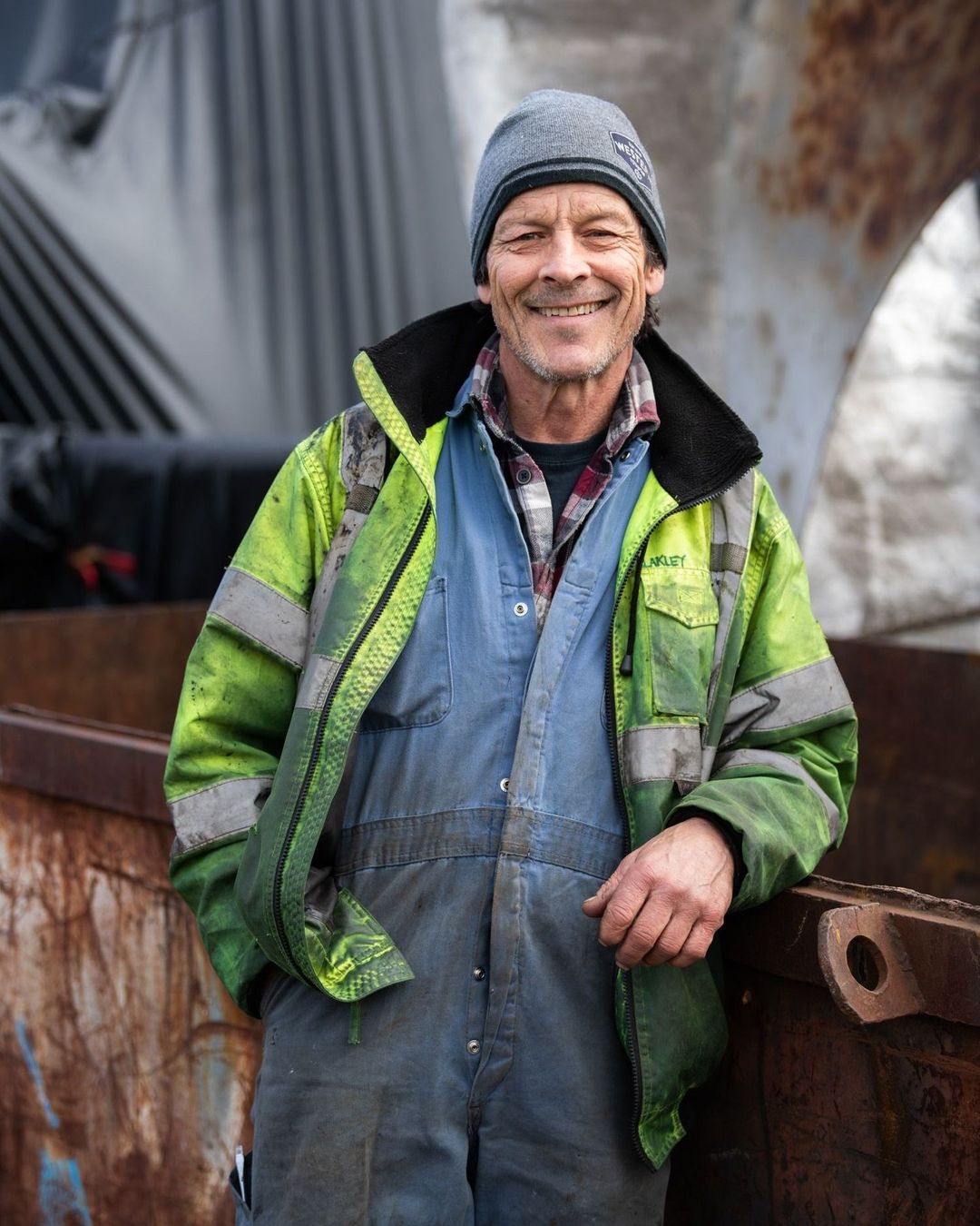 Smiling construction worker in blue overalls and a neon safety jacket standing in front of metal scrap, looking directly at the camera.