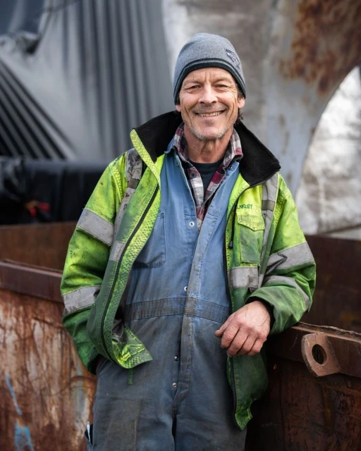 Smiling construction worker in blue overalls and a neon safety jacket standing in front of metal scrap, looking directly at the camera.