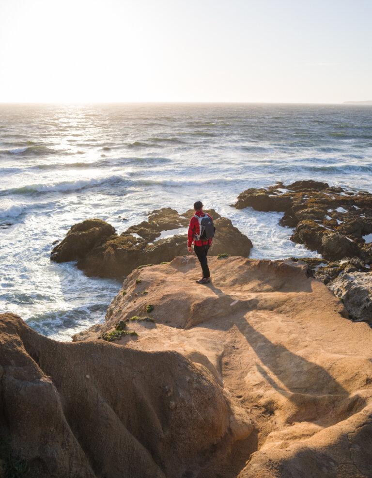 Adult male stands atop a rocky cliff overlooking the ocean in a red jacket and backpack. White waves crash against the rocks in sunlight.