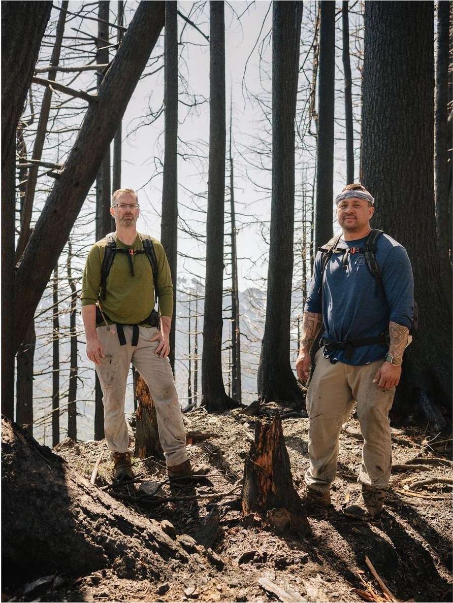 Two adult men standing in a forest with bare trees and a mountain in the background, wearing hiking clothes and carrying backpacks.