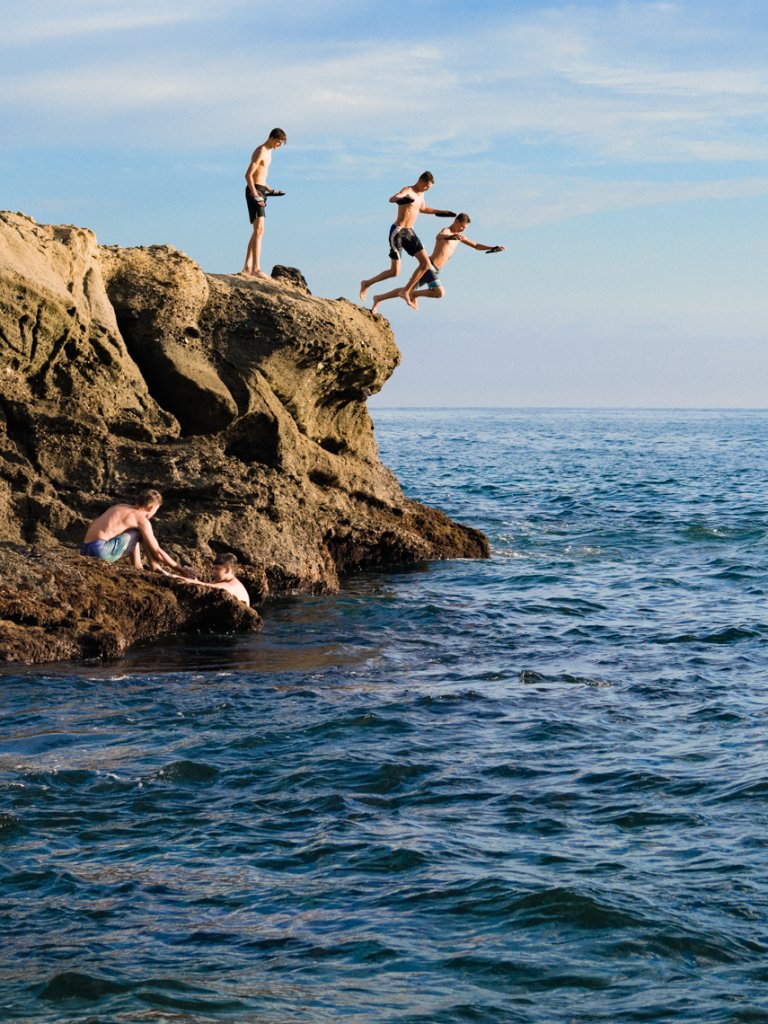 Group of young men jumping off rock cliff into ocean. Some are standing on the rocks, while others swim in the water below.