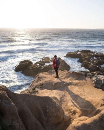 Man wearing a red jacket and backpack standing on a rocky coastal cliff overlooking the ocean at sunset by Peter Bohler