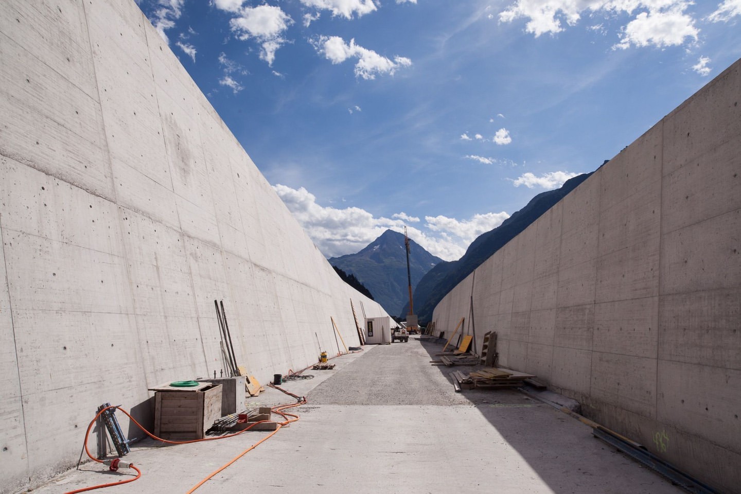 Concrete walls flank a road construction site with mountain views under a blue sky with clouds, utility boxes and hoses in foreground.
