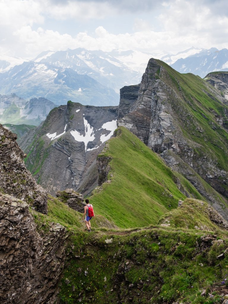 A lone hiker with a red backpack walks a grassy ridge with distant snowy mountains and dramatic rock formations under a cloudy sky.