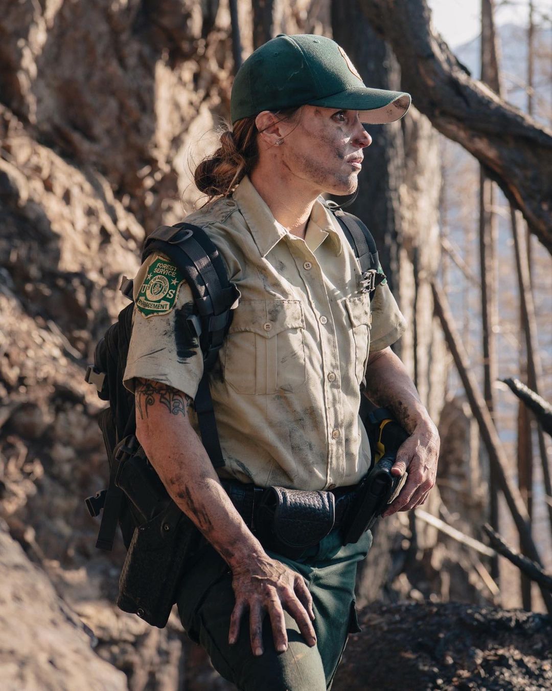 Adult female park ranger stands outdoors in uniform and baseball cap, looking off into the distance with forest and rocky landscape background.