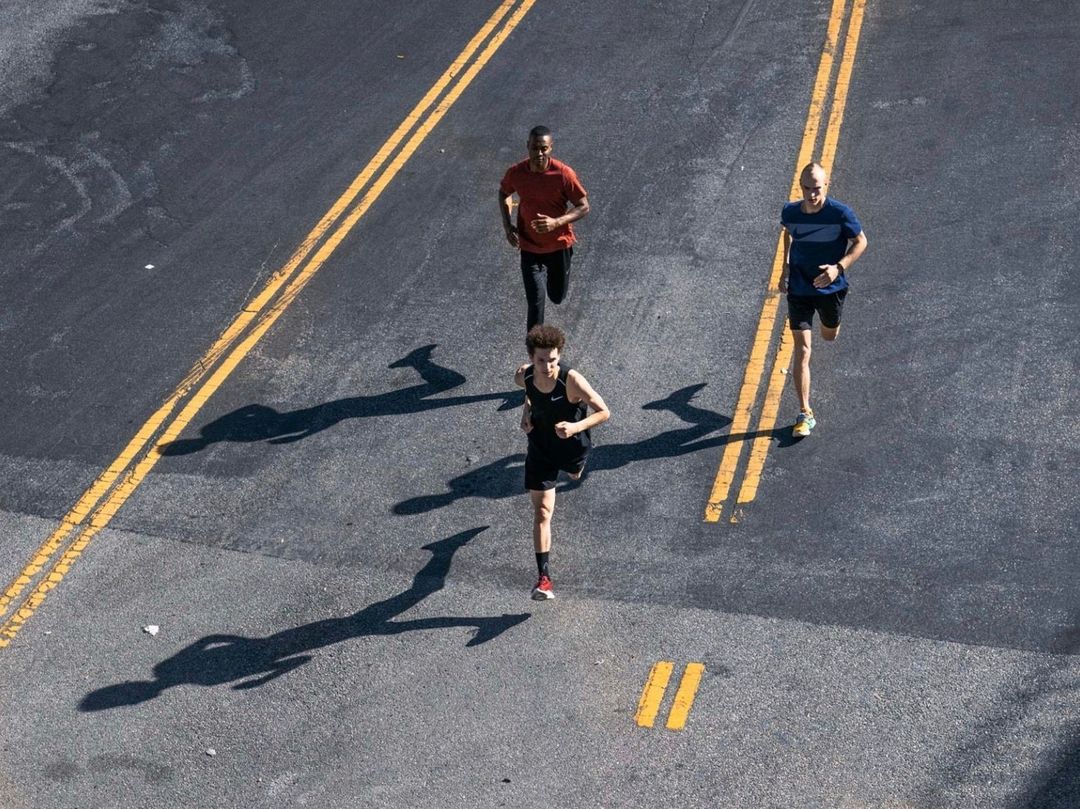 High-angle view of three adult athletes running along an asphalt road with yellow lane markings on a sunny day, casting shadows.