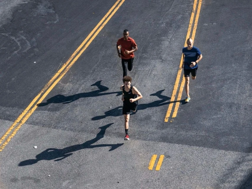 High-angle view of three adult athletes running along an asphalt road with yellow lane markings on a sunny day, casting shadows.