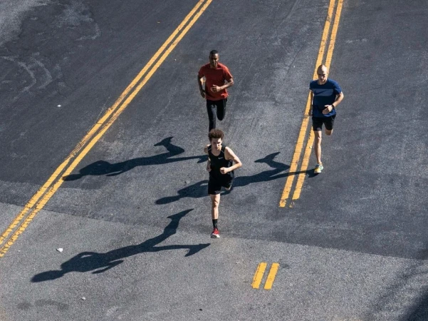High-angle view of three adult athletes running along an asphalt road with yellow lane markings on a sunny day, casting shadows.
