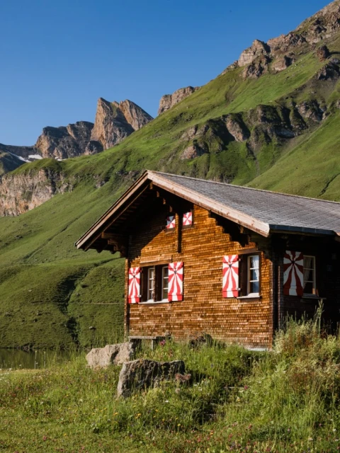 Wooden chalet nestled in a grassy valley with mountains under a clear blue sky. Red and white shutters decorate the windows.