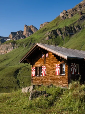 Wooden chalet nestled in a grassy valley with mountains under a clear blue sky. Red and white shutters decorate the windows.