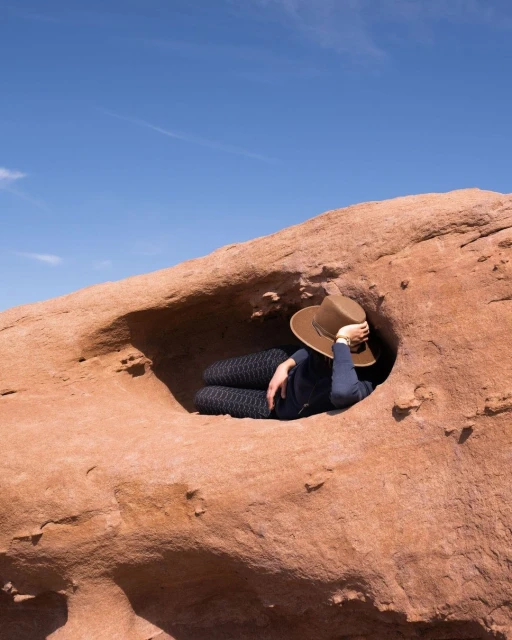 Woman rests in a sandstone rock formation with a hole, wearing a hat and patterned pants, under a clear blue sky, outdoors.