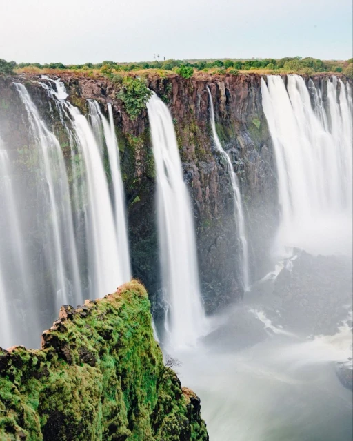 Dramatic, cascading waterfalls featuring long exposure motion, lush greenery, and rugged cliffs under an overcast sky, capturing nature's raw power.