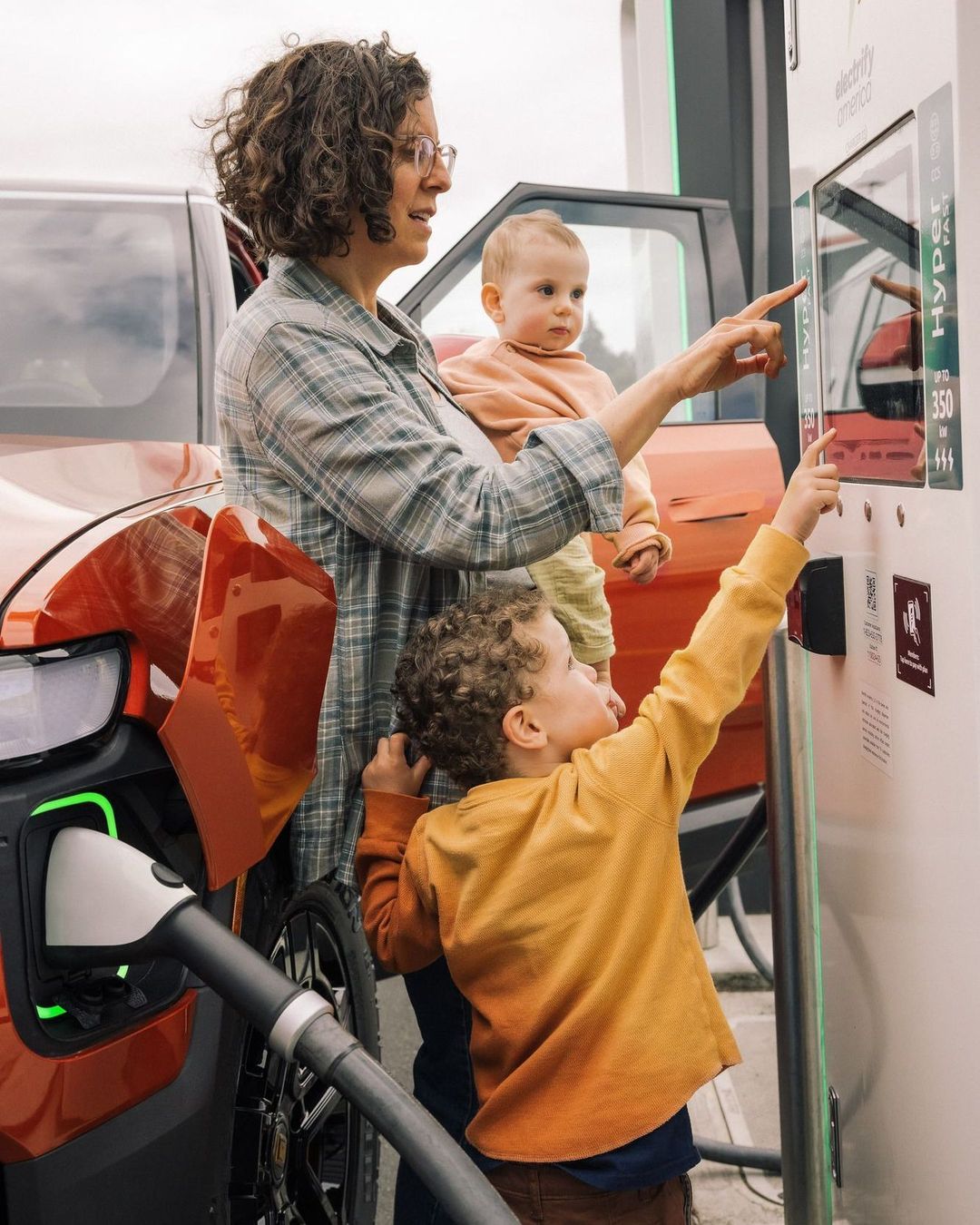 A woman and two children interact with an electric car charging station. The orange vehicle is plugged in, while the family examines the display.