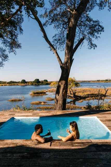 A mixed-race couple relaxes in a pool at the edge of a river on a sunny day, enjoying drinks and the natural landscape.