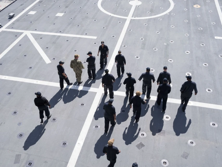 Overhead shot of a group of Navy personnel in black uniforms standing on the grey deck of a ship marked with white lines and circles.