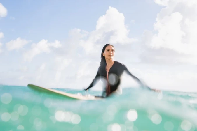 Woman in wetsuit paddles surfboard in the ocean. Turquoise water and cloudy blue skies complete the scene on a sunny day.