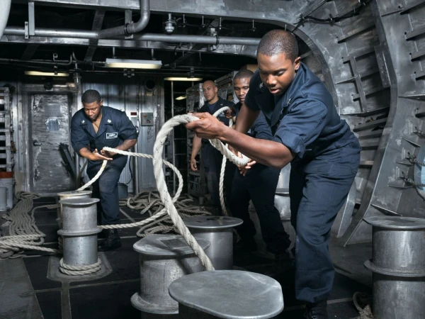 Four sailors in navy uniforms handle thick white ropes on a US Navy ship, working together to secure or manage the lines.