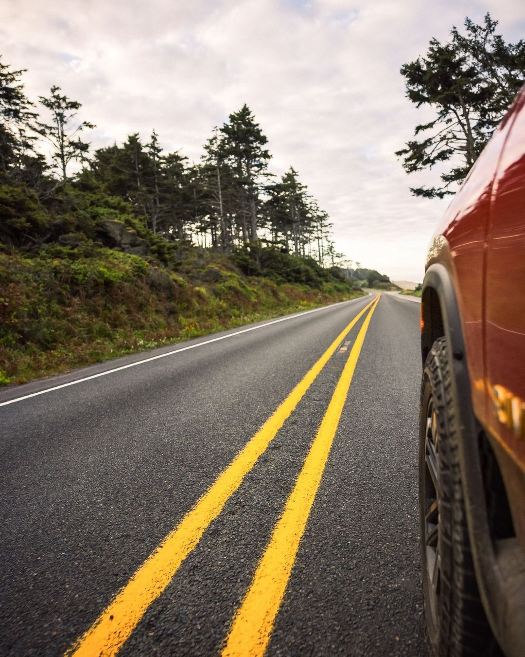 A red truck drives down a long highway road lined with trees under a cloudy sky, captured from a low angle perspective.