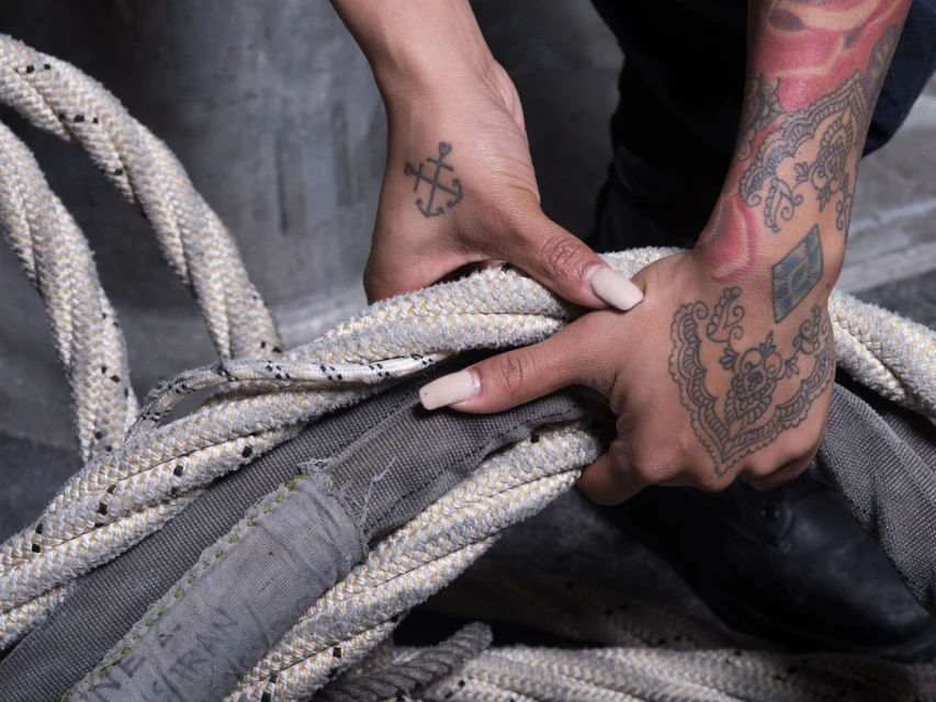 Close-up on tattooed hands holding a pile of grey and white ropes, set against a dark concrete backdrop, showcasing texture and detail.
