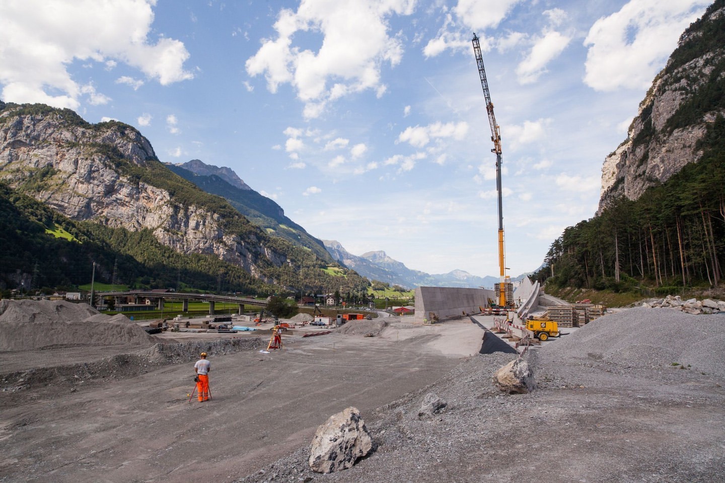 Construction site amid Swiss Alps features workers, crane, gravel piles, and bridge foundation on a partly cloudy day near mountains.