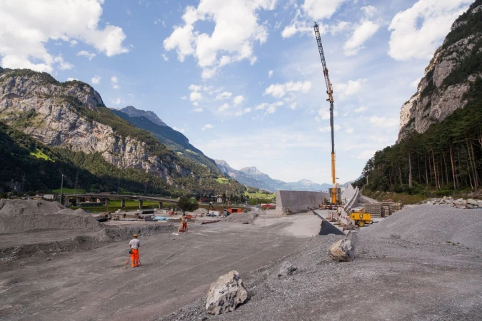 Construction site amid Swiss Alps features workers, crane, gravel piles, and bridge foundation on a partly cloudy day near mountains.