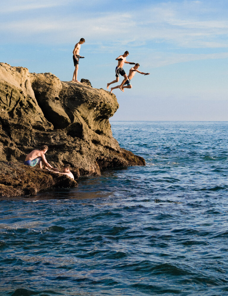Several shirtless men jumping off rocky cliff into ocean against blue sky. Cliff diving and summertime fun in outdoor environment.