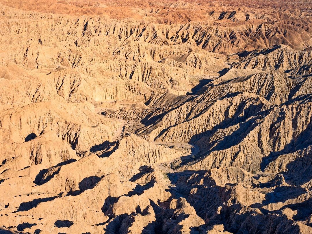 Aerial view of eroded badlands landscape in sunlight, revealing textures and patterns of the arid terrain with hills and valleys.