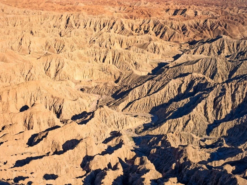 Aerial view of eroded badlands landscape in sunlight, revealing textures and patterns of the arid terrain with hills and valleys.