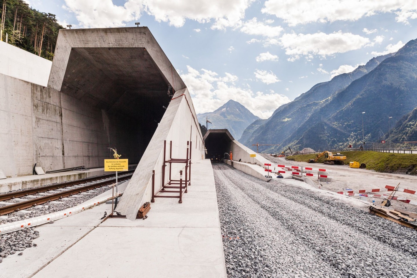Tunnel construction site with concrete entrances, railway tracks, gravel, construction equipment, mountains, and a blue sky with clouds.