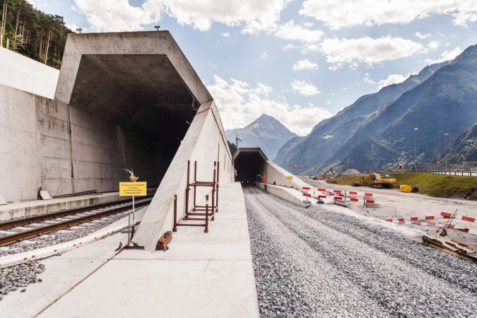 Tunnel construction site with concrete entrances, railway tracks, gravel, construction equipment, mountains, and a blue sky with clouds.