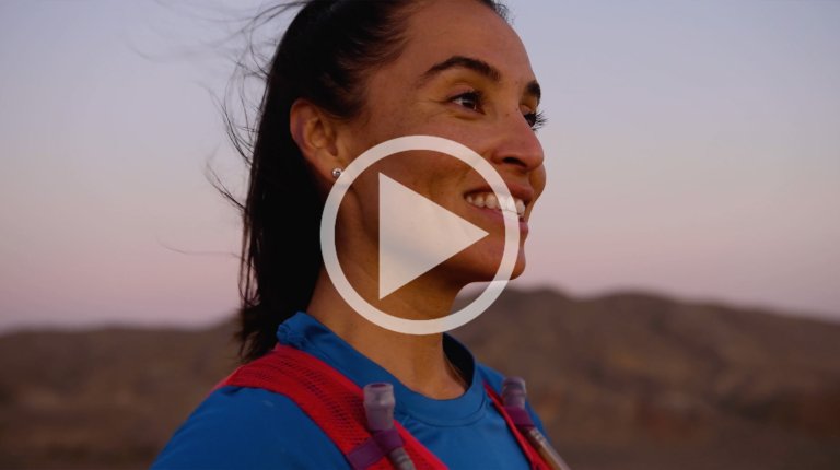 Adult woman smiles outdoors in a blue athletic shirt with a hydration pack, against a backdrop of blurred mountains at sunset.