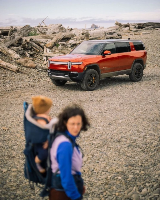 An orange Rivian SUV is parked on a rocky beach near driftwood, with a woman and child in a carrier in the foreground.