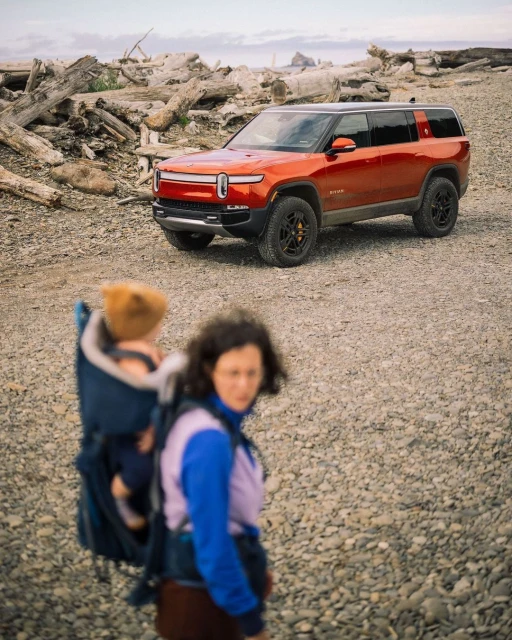 An orange Rivian SUV is parked on a rocky beach, with a woman carrying a baby in a carrier walking towards the camera.