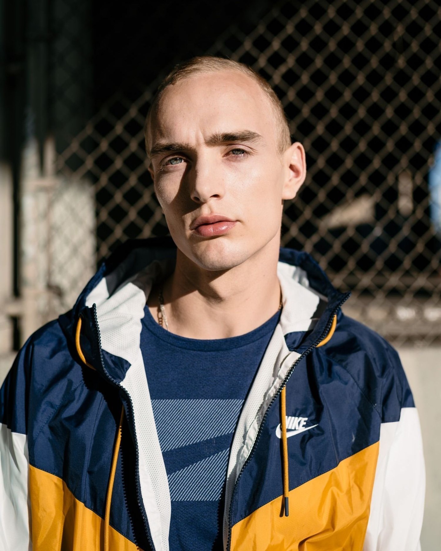 Portrait of a young adult man wearing a Nike jacket, facing forward, with a chain-link fence and light and shadows in the background.