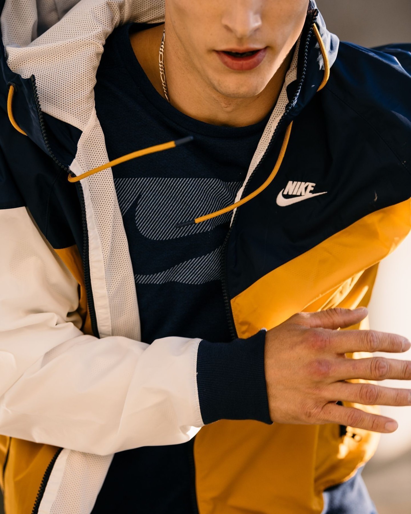 Close-up of a young adult man wearing a black T-shirt and a color-blocked Nike windbreaker jacket in white, gold, and black.