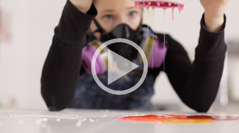 Young woman with a respirator pours pink liquid from a strainer, creating a vibrant splatter on a white surface, indoor, eye-level.
