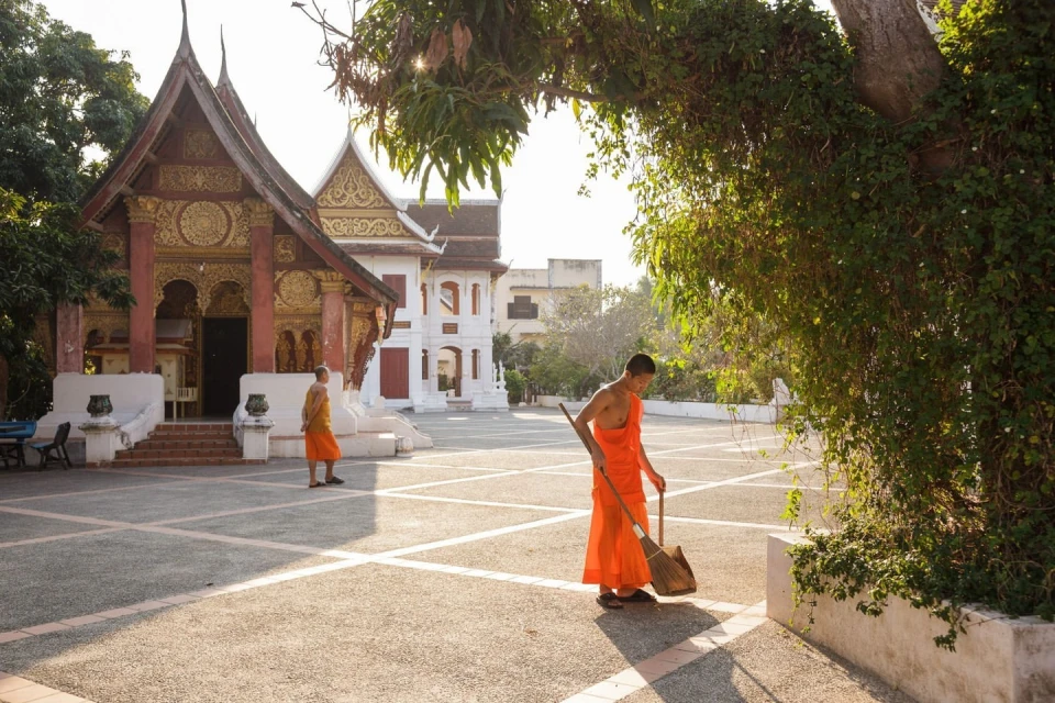 Two monks in orange robes sweeping the courtyard of a temple complex with traditional architecture and lush trees on a sunny day.