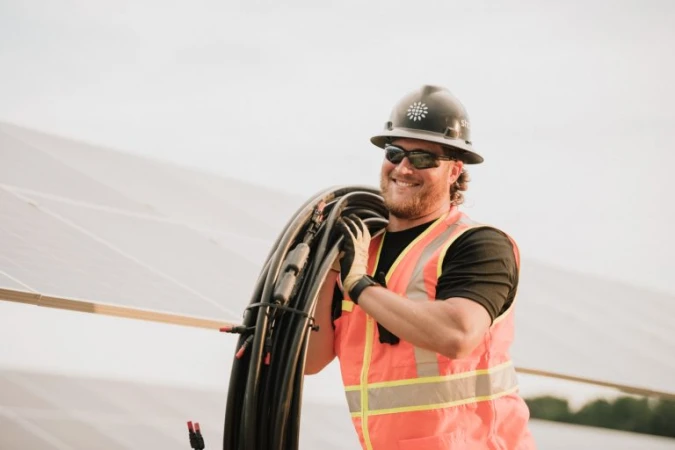Smiling construction worker wearing hard hat and safety vest holding black cables on a sunny day, standing by solar panels.