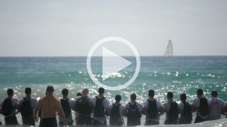 Group of men standing in the ocean surf facing away from the camera toward a distant sailboat on the horizon