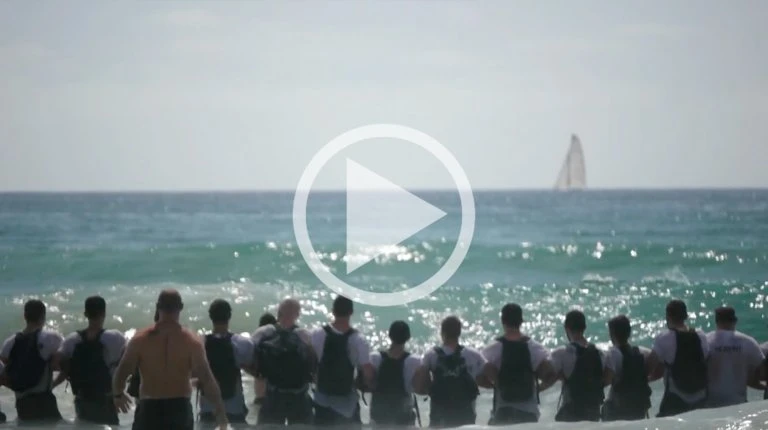 Group of men standing in the ocean surf facing away from the camera toward a distant sailboat on the horizon