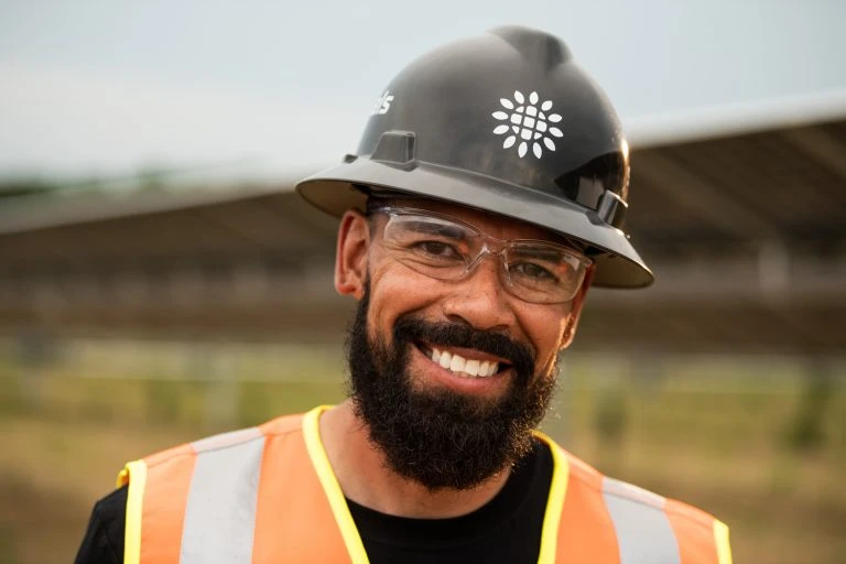 Smiling man in hardhat and safety vest stands outdoors; worker with beard and safety glasses looks directly at the camera.